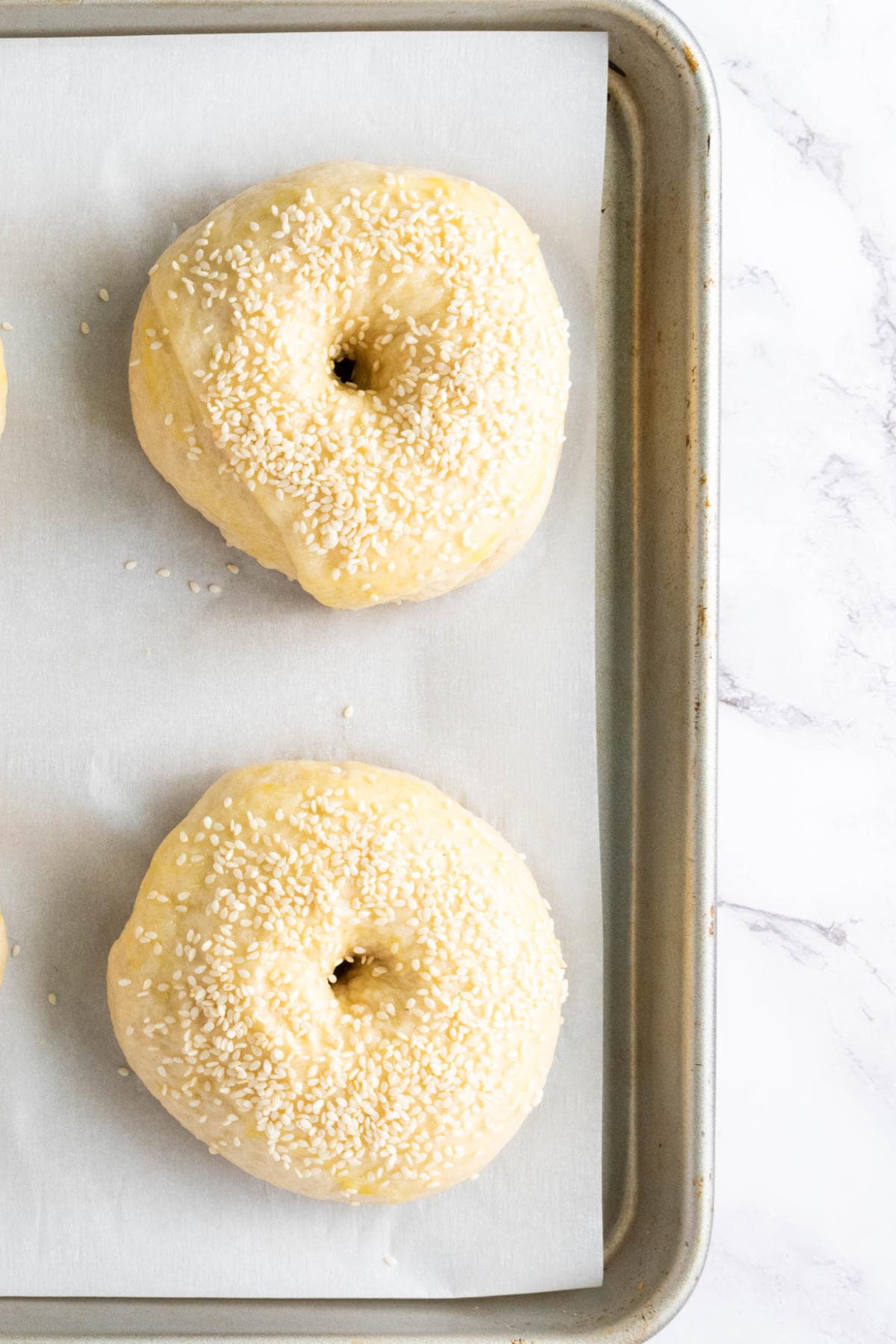 Two unbaked bagels topped with sesame seeds sit on a parchment-lined baking sheet, ready to be baked. The image is shot from above on a light marble surface.