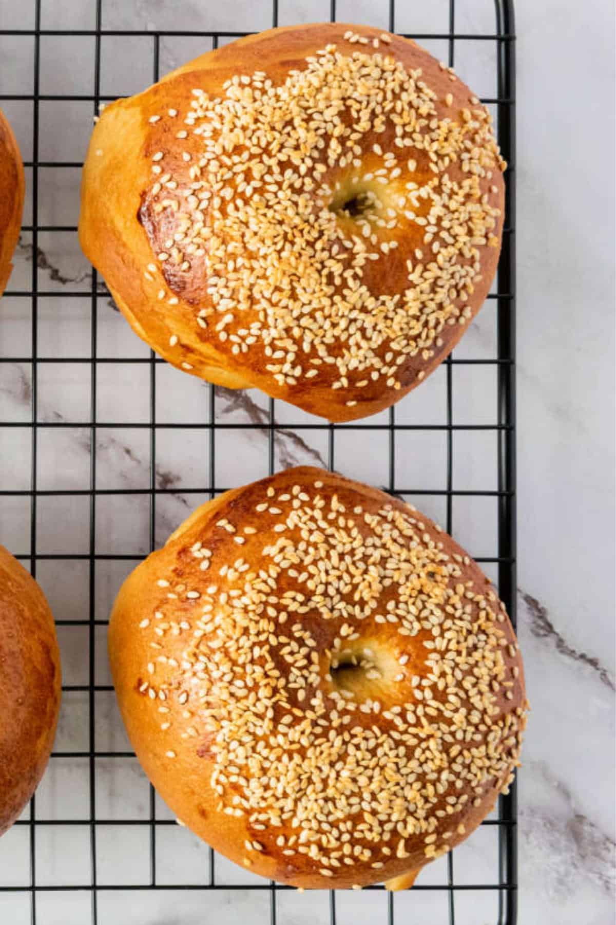 Two golden brown bagels topped with sesame seeds are cooling on a black wire rack, placed on a white marble surface.