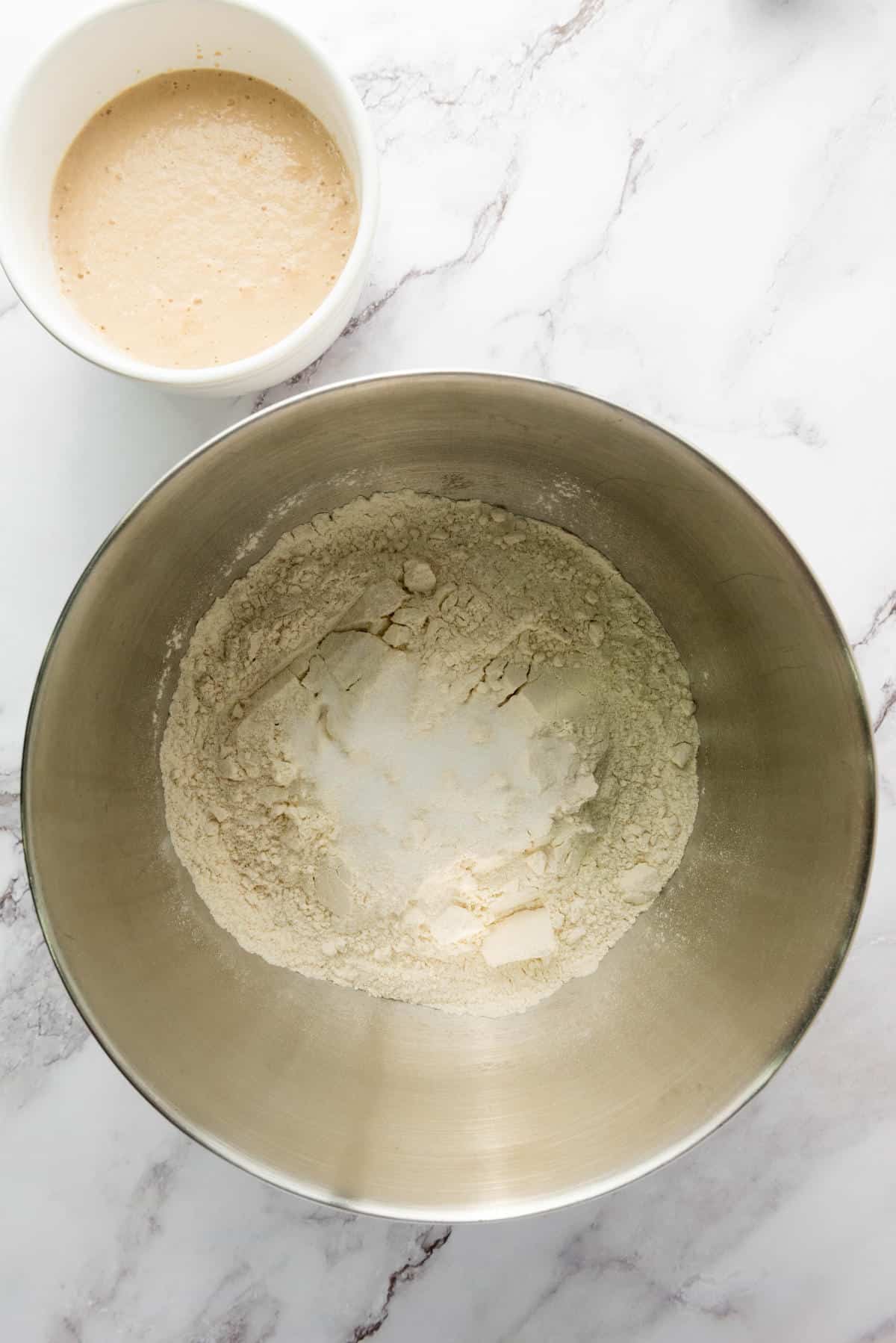 A metal mixing bowl with flour and other dry ingredients sits on a marble countertop next to a small white bowl containing a beige liquid mixture.