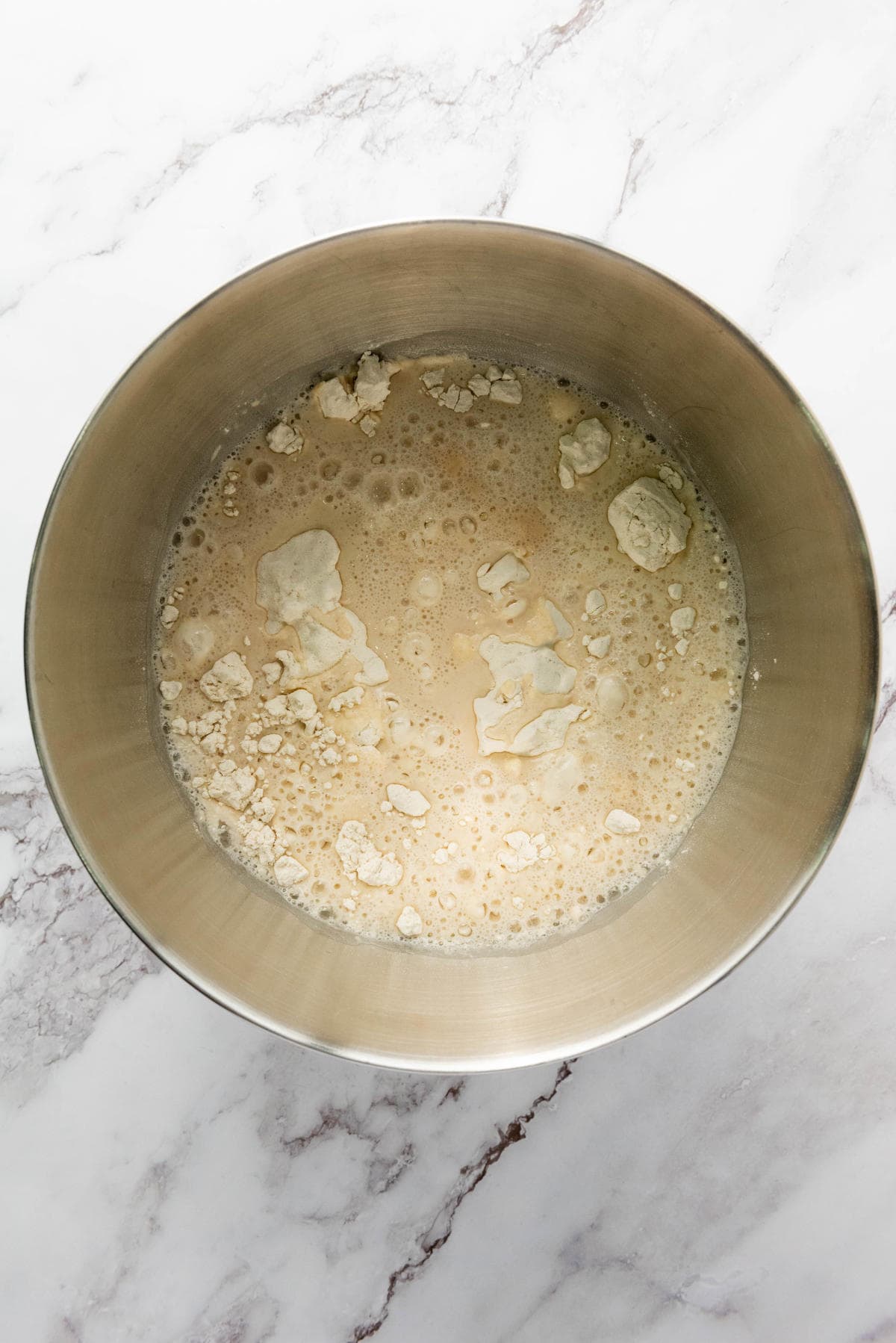 A metal mixing bowl containing a mixture of flour, yeast, and water sitting on a white marble countertop. Some flour is floating on top of the liquid.