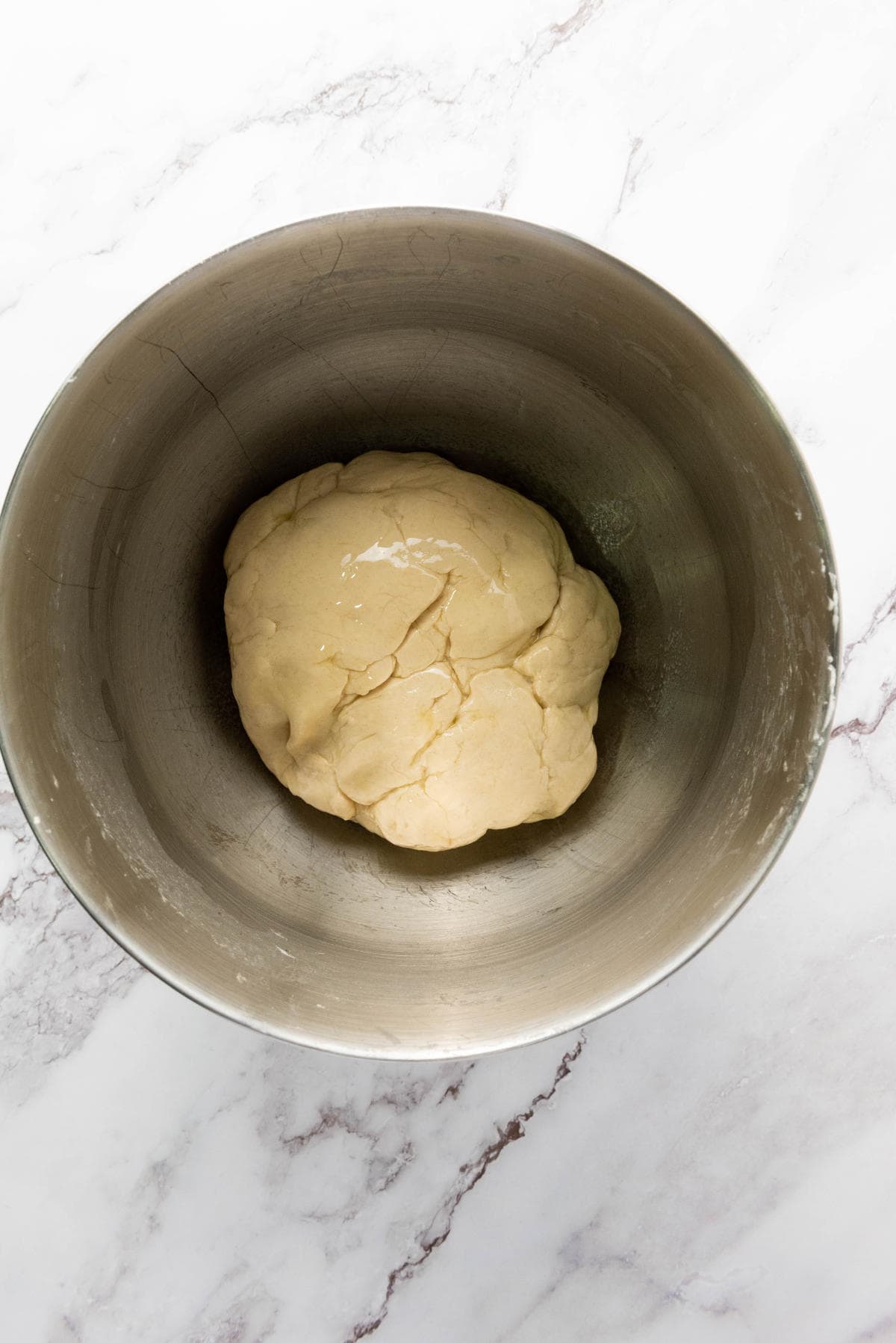 A ball of dough sits in the center of a large metal mixing bowl on a white marble countertop.