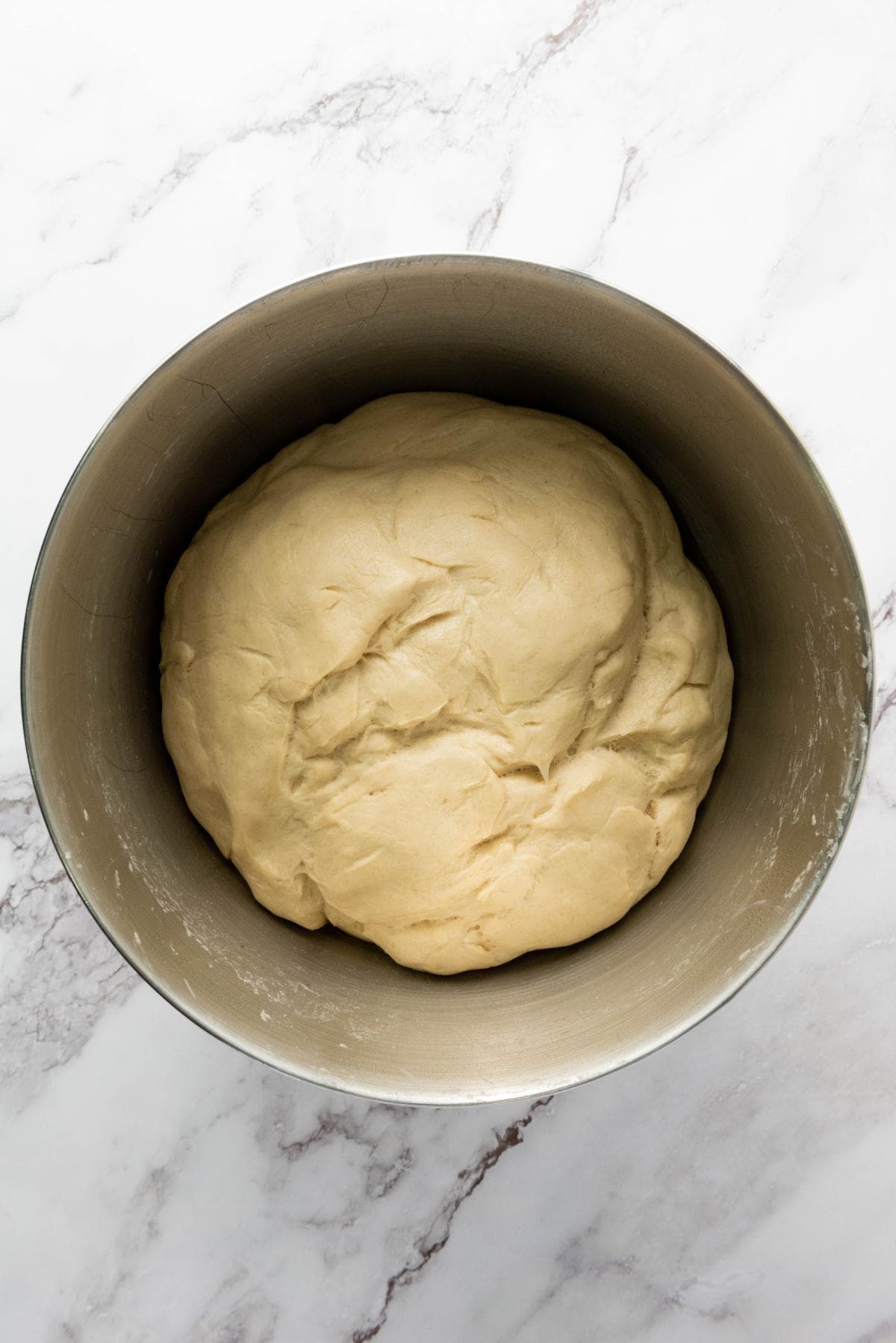 A ball of smooth, risen dough sits in a large metal mixing bowl on a white marble countertop.