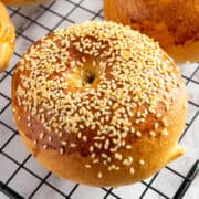 A golden-brown bagel topped with sesame seeds sits on a black wire cooling rack. The bagel has a shiny surface and a slightly raised, rounded shape.
