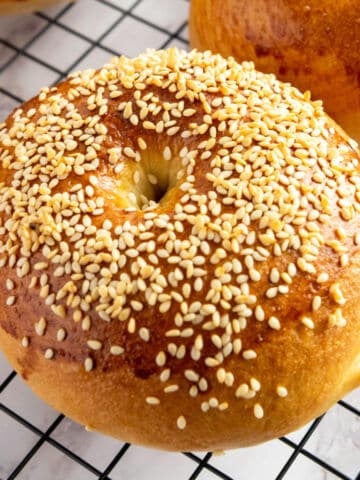 A golden-brown bagel topped with sesame seeds sits on a black wire cooling rack. The bagel has a shiny surface and a slightly raised, rounded shape.