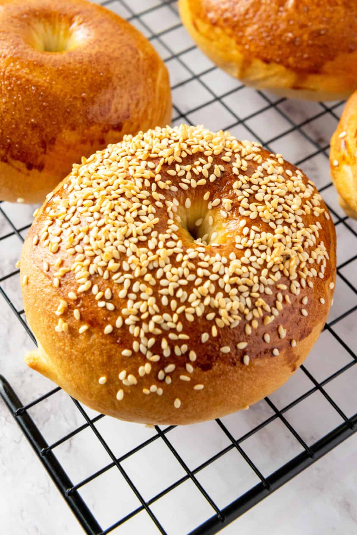 A golden brown bagel topped with sesame seeds rests on a black wire cooling rack, surrounded by other plain bagels.