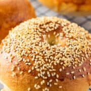 A close-up of a golden brown bagel topped with sesame seeds, resting on a wire cooling rack. Other bagels are visible in the background.