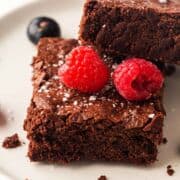 A close-up of two chocolate brownies stacked on a plate, topped with two fresh raspberries and a sprinkle of flaky sea salt. A blueberry sits in the background.