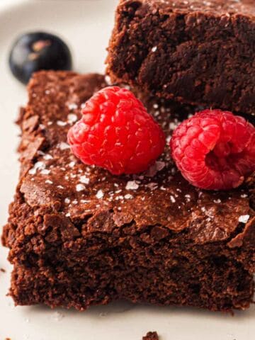 A close-up of two chocolate brownies stacked on a plate, topped with two fresh raspberries and a sprinkle of flaky sea salt. A blueberry sits in the background.