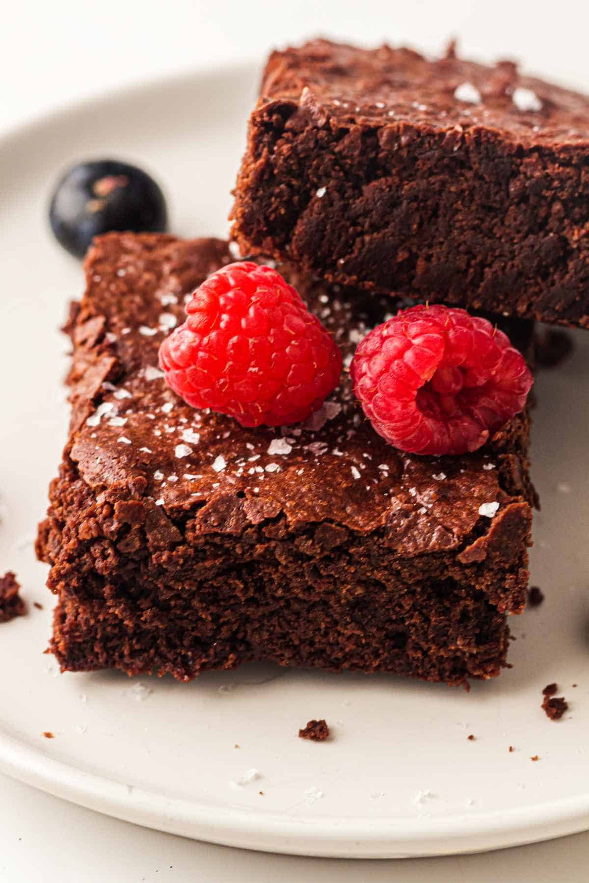 Two fudgy sheet pan brownies topped with raspberries and flaky sea salt on a white plate.
