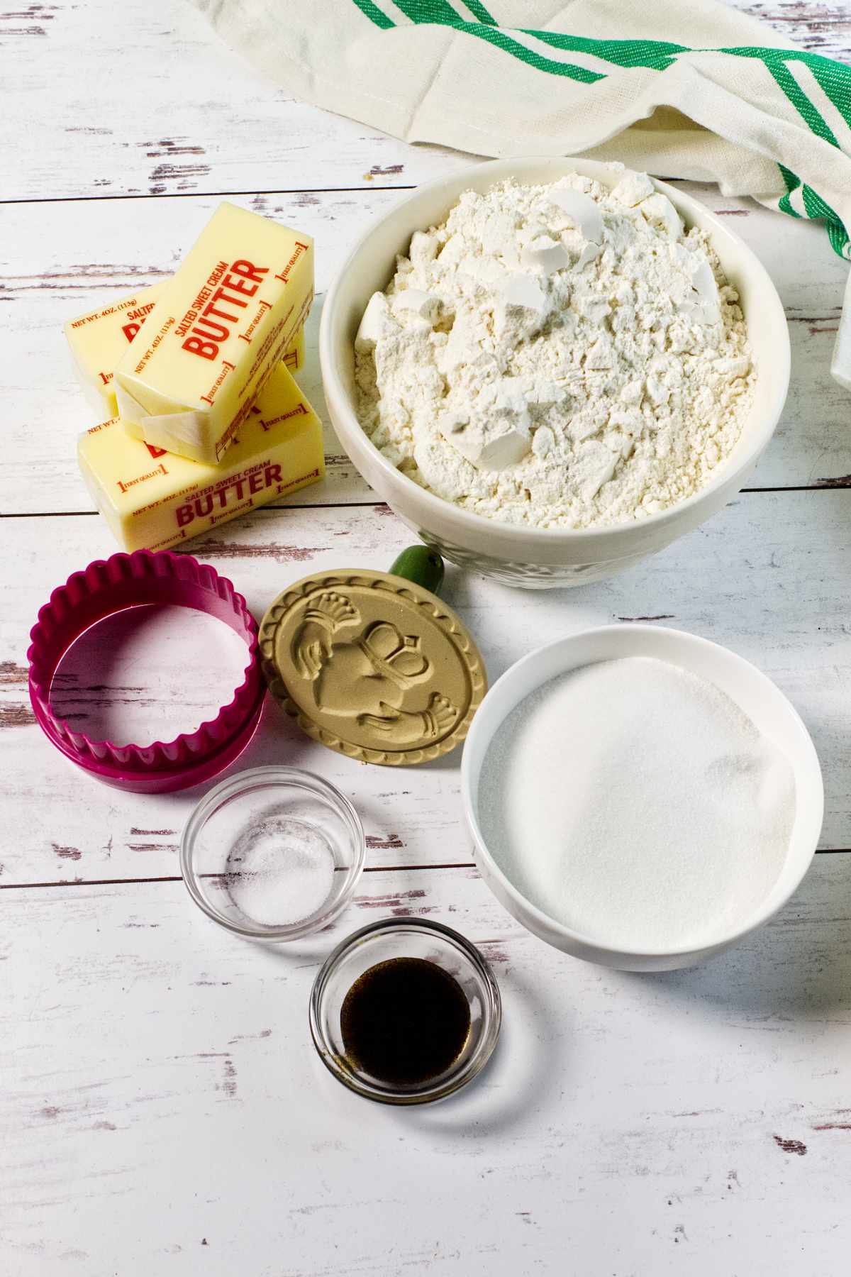 A bowl of flour, a bowl of sugar, three sticks of butter, two small bowls with vanilla extract and salt, a green and gold cookie stamp for stamped shortbread cookies recipe, and a round biscuit cutter on a white wooden table with a striped towel.
