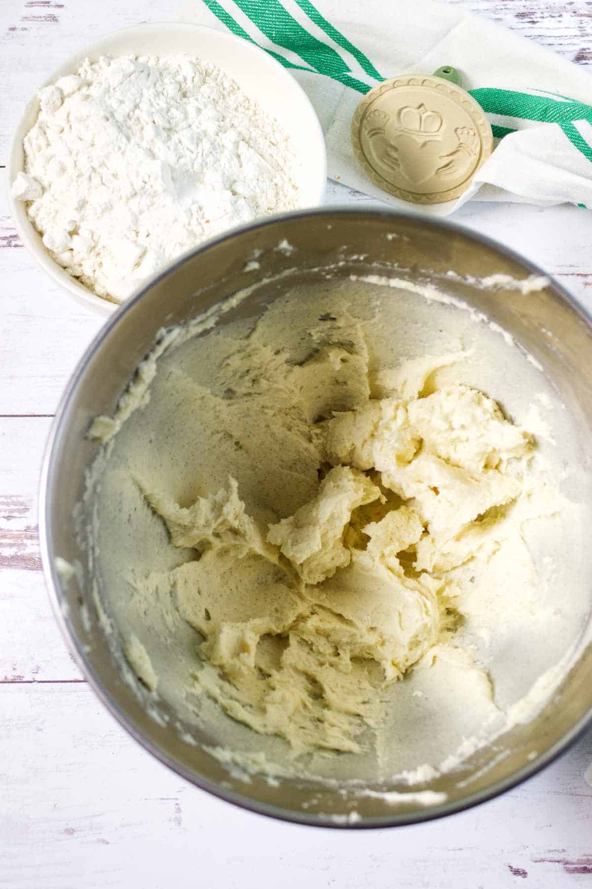 A metal mixing bowl with creamed dough, ready for cookies, sits beside a bowl of flour on a white surface. A green-striped kitchen towel and a wooden cookie stamp are in the background.
