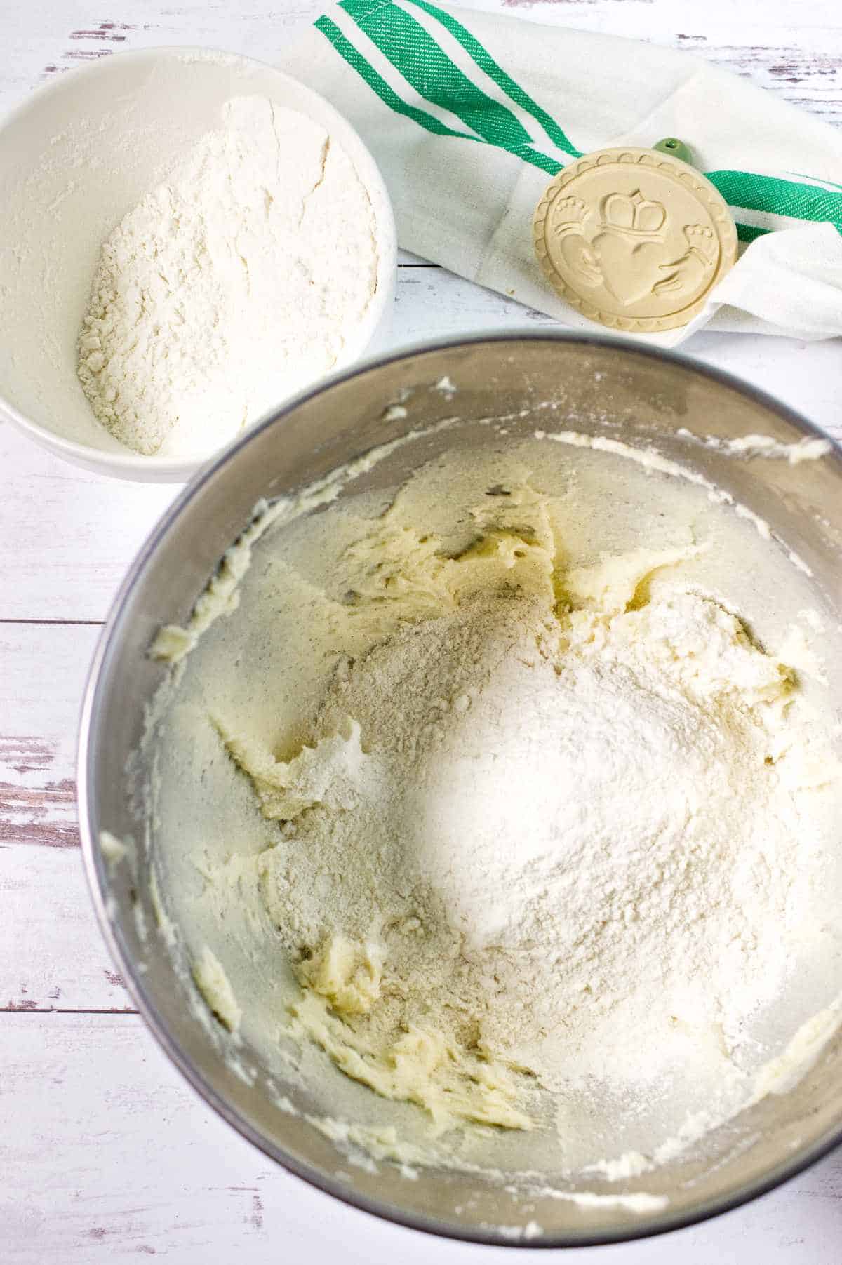 A metal mixing bowl with creamed dough and flour being added, perfect for making shortbread, sits next to a white bowl of flour, a green-striped towel, and a wooden cookie stamp on a white wooden surface.