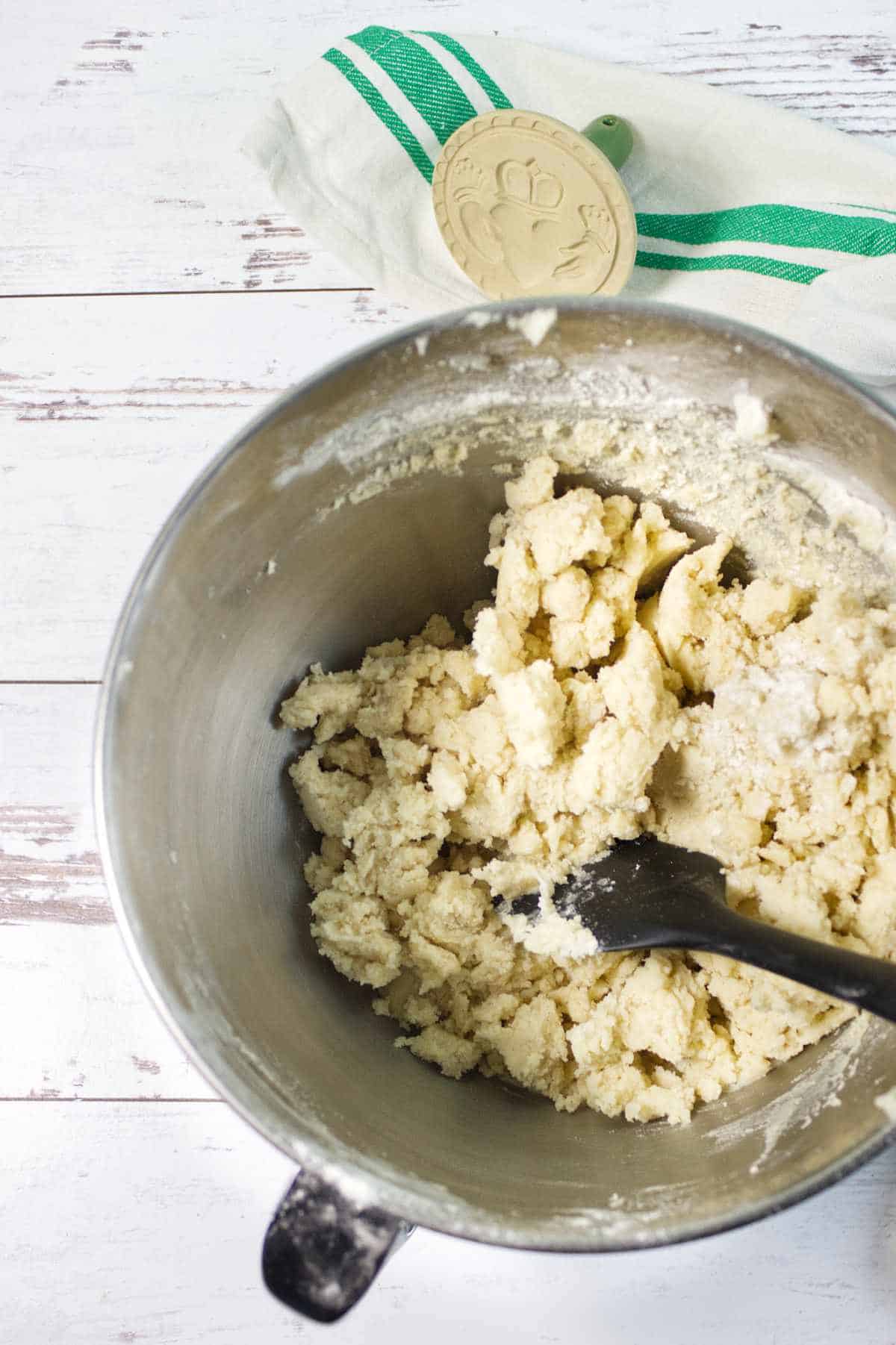 A metal mixing bowl filled with crumbly dough for cookies sits on a white wooden surface, with a black spatula inside. In the background, a tea towel and a beige cookie press are visible.