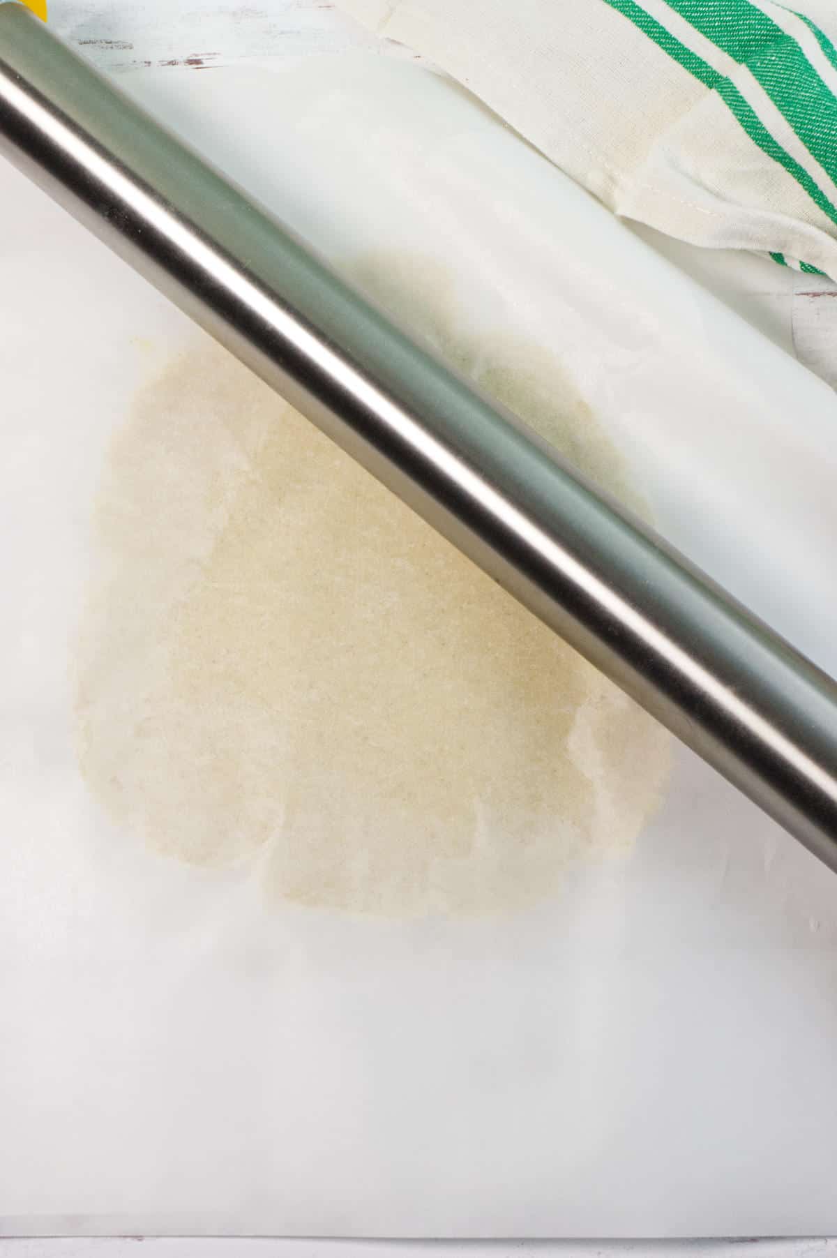 A metal rolling pin presses down on parchment paper with dough underneath, partially flattened, with a green-striped cloth in the background.