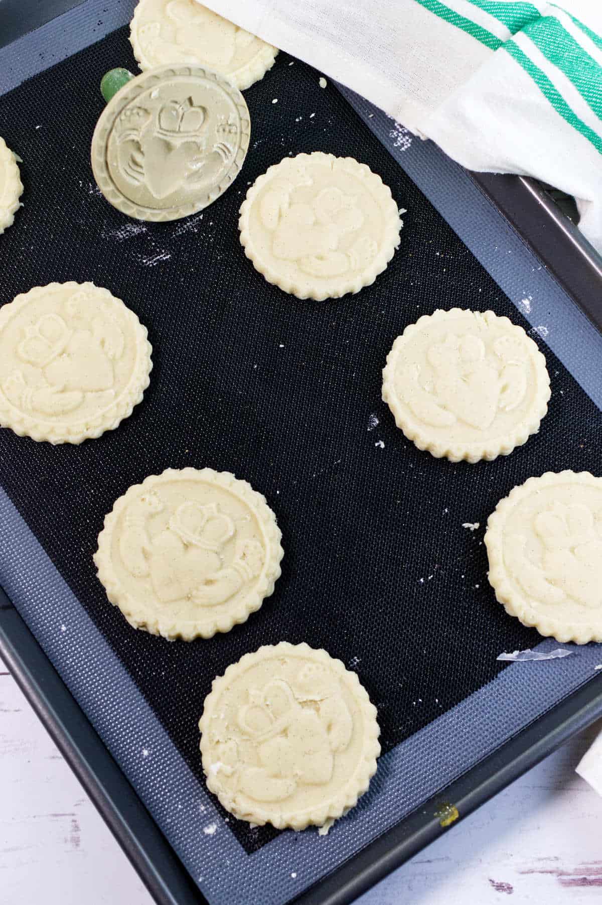 A baking sheet with several round, stamped shortbread cookies and a decorative cookie stamp nearby. The cookies are evenly spaced and have a floral pattern. A green and white towel is partially visible in the corner.