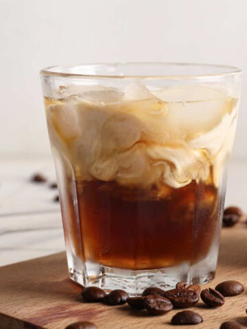 A glass of iced coffee with milk swirling through it sits on a wooden board, surrounded by coffee beans, evoking the rich, creamy look of a white Russian cocktail against a blurred white background.