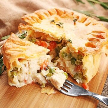 A golden, flaky chicken pot pie on a wooden board, with a slice cut out revealing chicken, peas, and carrots inside. A fork holds a bite of filling in front of the pie. Fresh rosemary is in the background.