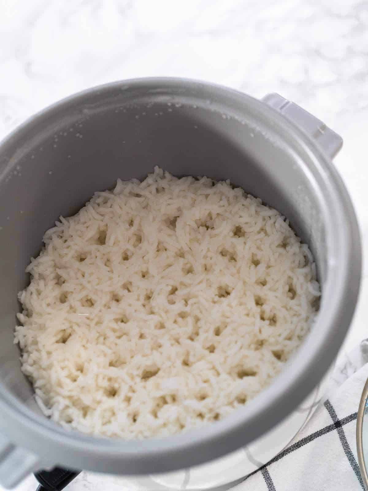 Cooked white rice inside a rice cooker, with the lid open and the fluffy grains visible. The rice cooker sits on a white surface with a checkered cloth partially visible nearby.