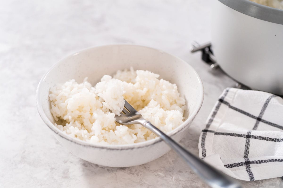 A white bowl filled with cooked white rice and a fork resting inside, next to a checkered napkin and part of a rice cooker on a light-colored surface.
