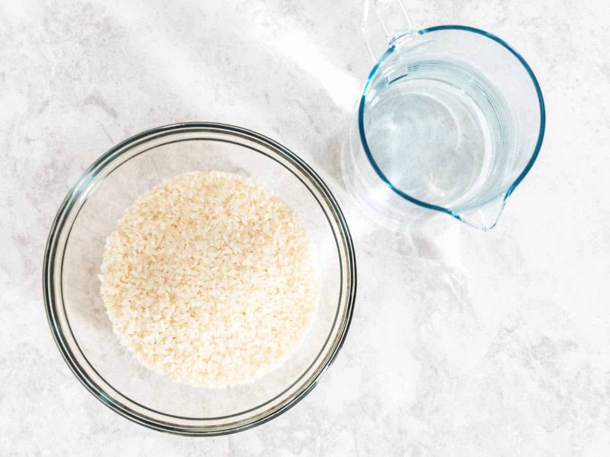 A glass bowl filled with uncooked white rice sits next to a clear measuring cup of water on a light-colored countertop.