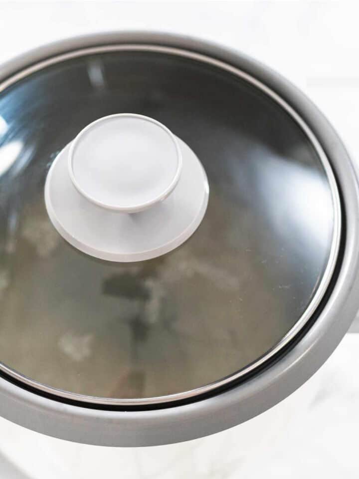 A close-up view of a glass rice cooker pot lid with a white handle, resting on a gray cooking pot. The lid reflects some light and the surrounding area.