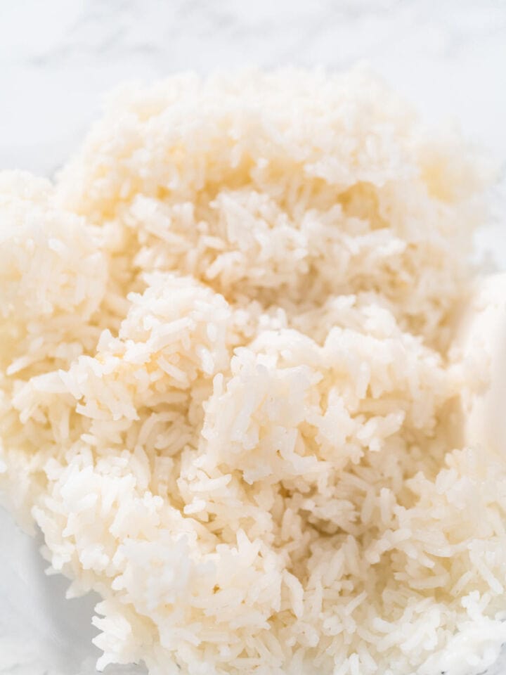 A close-up of a mound of cooked white rice, with individual grains visible and a soft, fluffy texture. The background is blurred and light in color.