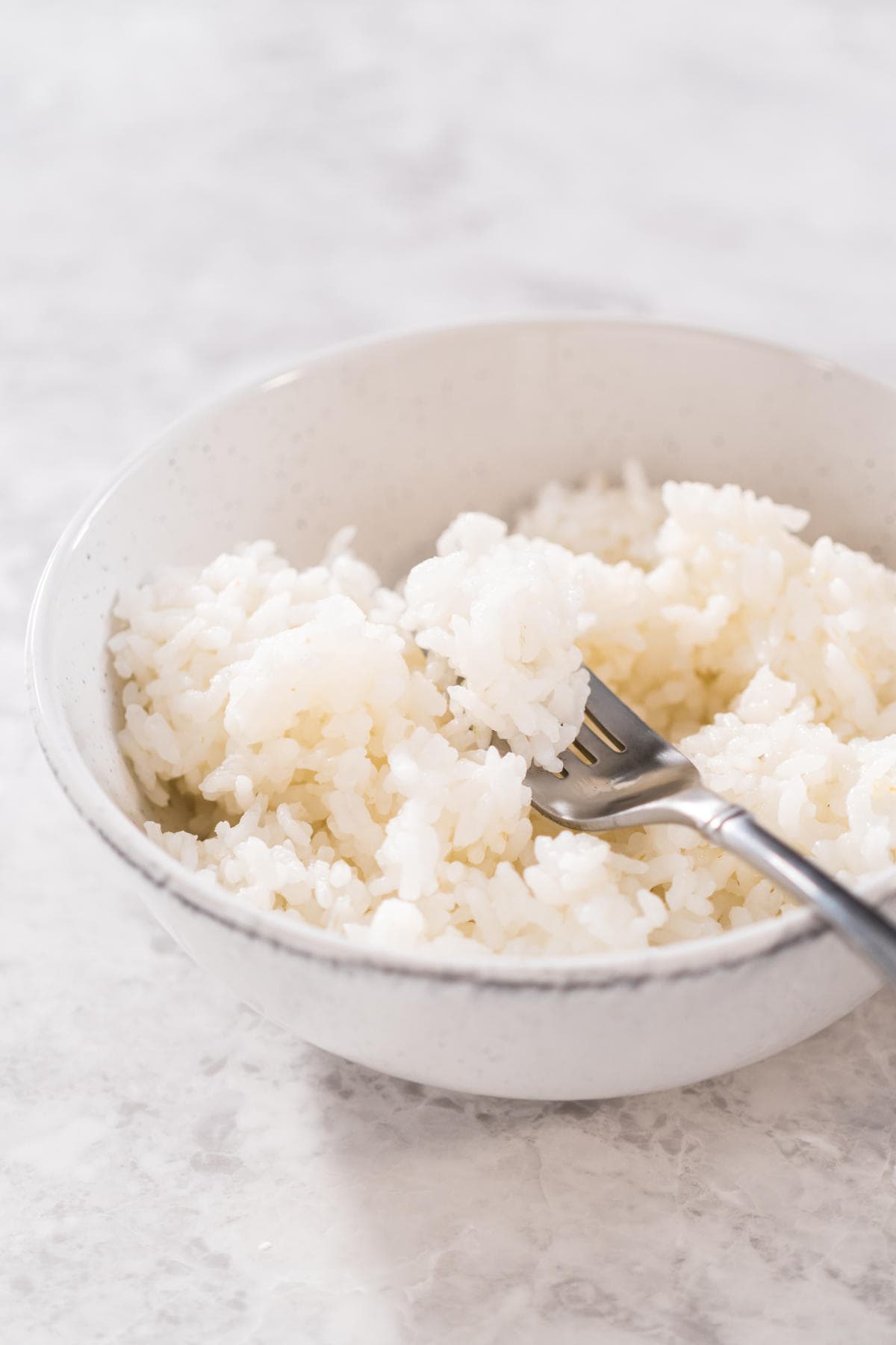 A white bowl filled with cooked white rice sits on a marble surface, with a metal fork resting inside the bowl.