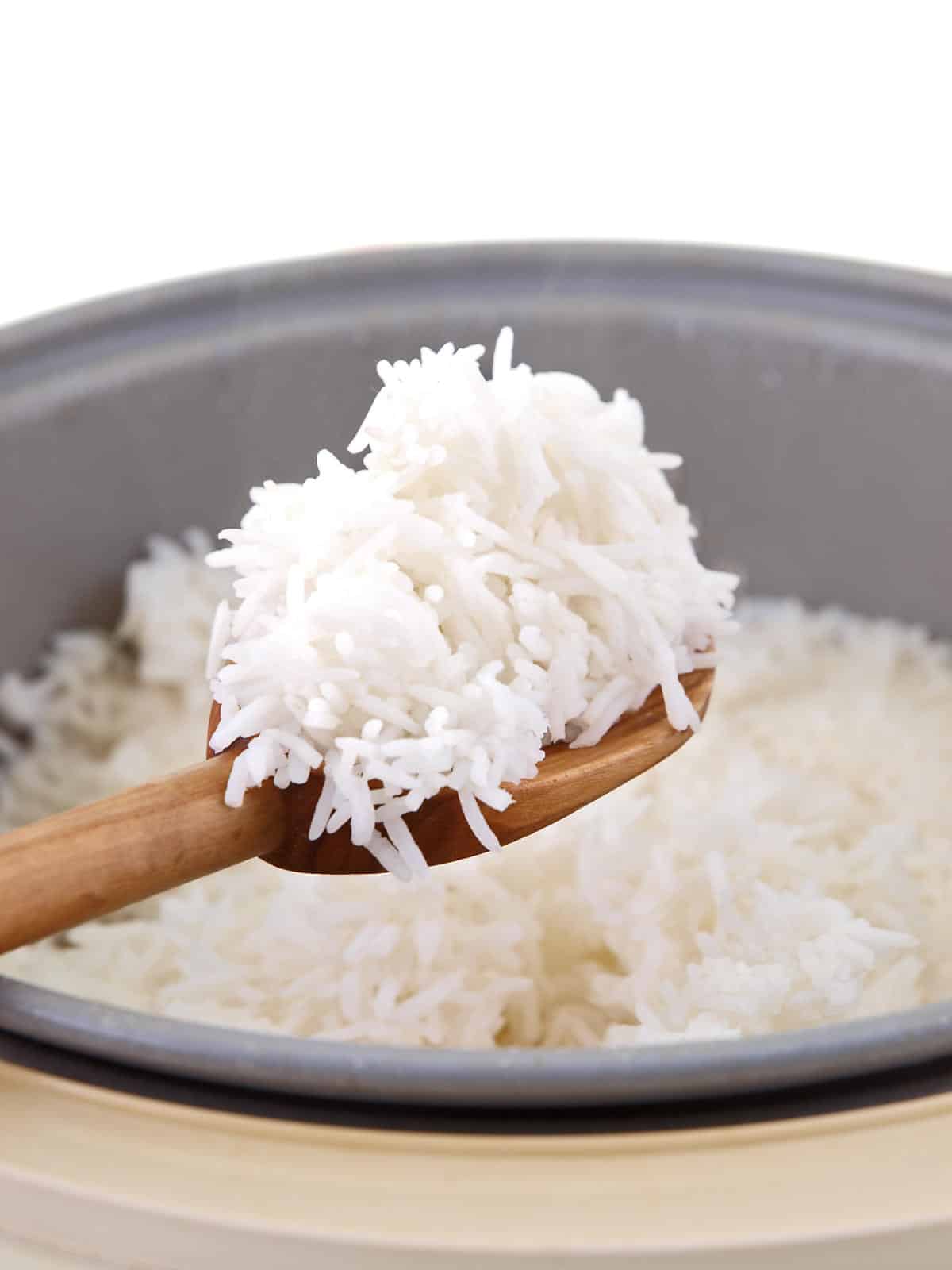A wooden spoon holding a serving of cooked white rice above a rice cooker filled with more rice.