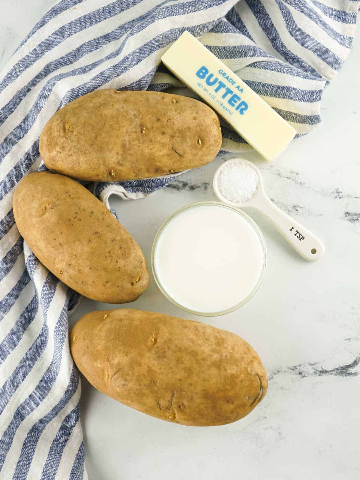 Ingredients for copycat Bob Evans mashed potatoes: russet potatoes, butter, milk, and salt on a white surface with a blue striped cloth.