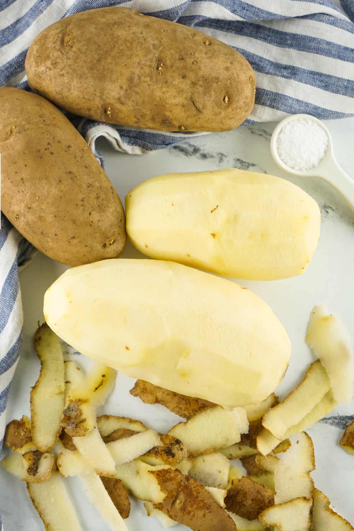Whole and peeled russet potatoes with potato peels on a white marble surface while preparing copycat Bob Evans mashed potatoes.