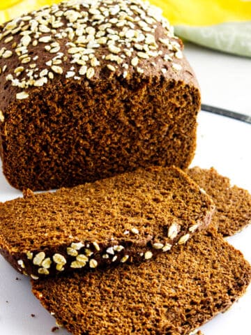 A loaf of dark, whole-grain bread topped with oats sits on a white surface, with two slices cut and lying in front of it. The bread looks dense and moist, with a rustic homemade appearance.