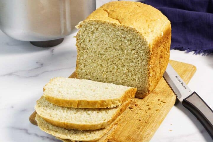 A loaf of homemade oatmeal bread sits on a wooden cutting board with two slices cut. A bread knife lies next to the bread, and a kitchen towel and metallic appliance are in the background.