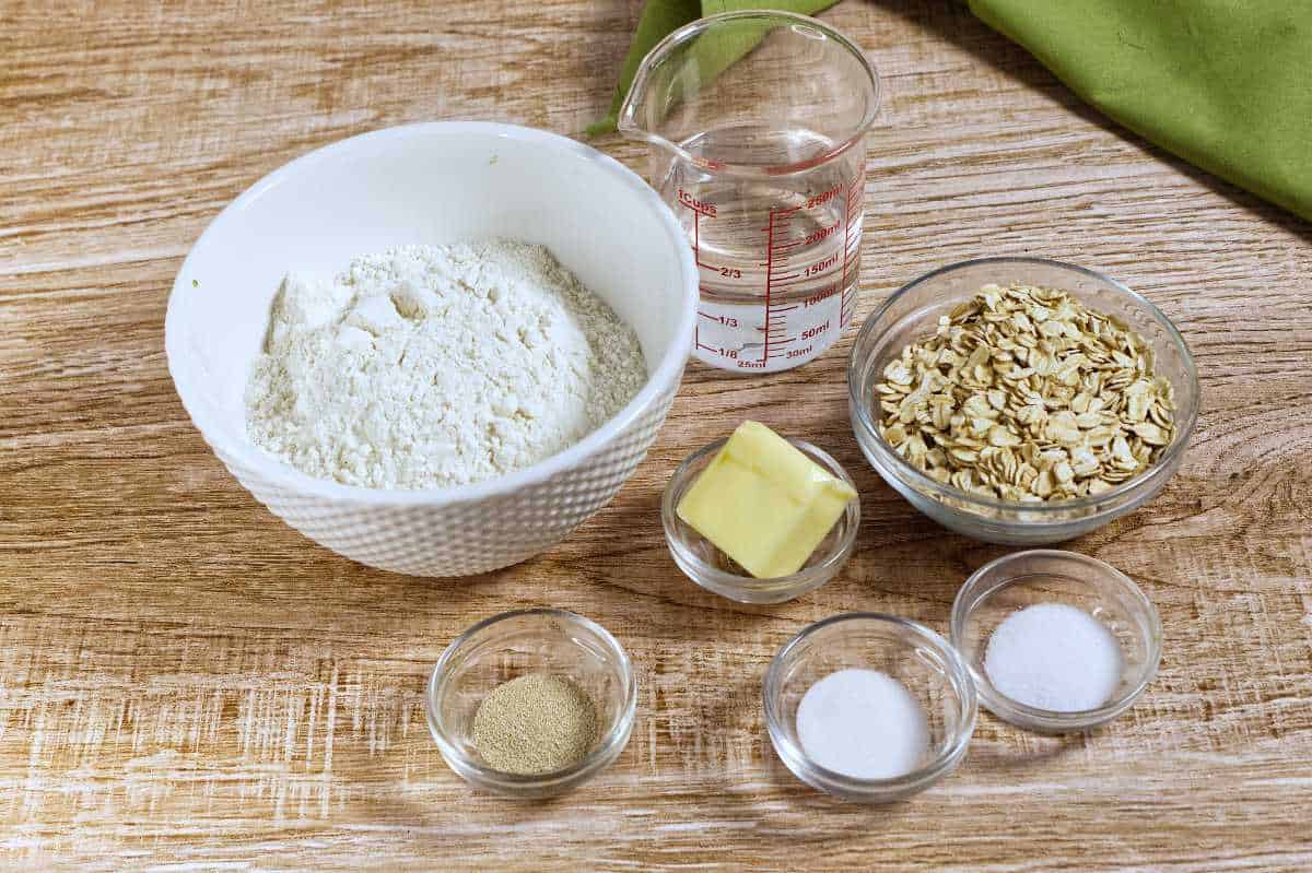 A bowl of flour, a glass measuring cup of water, a bowl of rolled oats, a pat of butter, and small bowls of yeast, sugar, and salt arranged on a wooden surface.