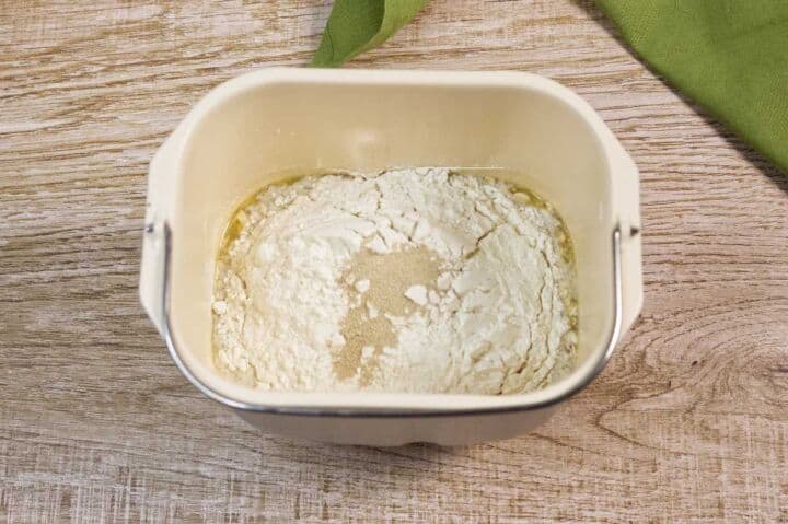 A bread machine pan filled with flour, yeast, oil, and water sits on a wooden surface next to a green cloth. The ingredients are unmixed and ready for baking.