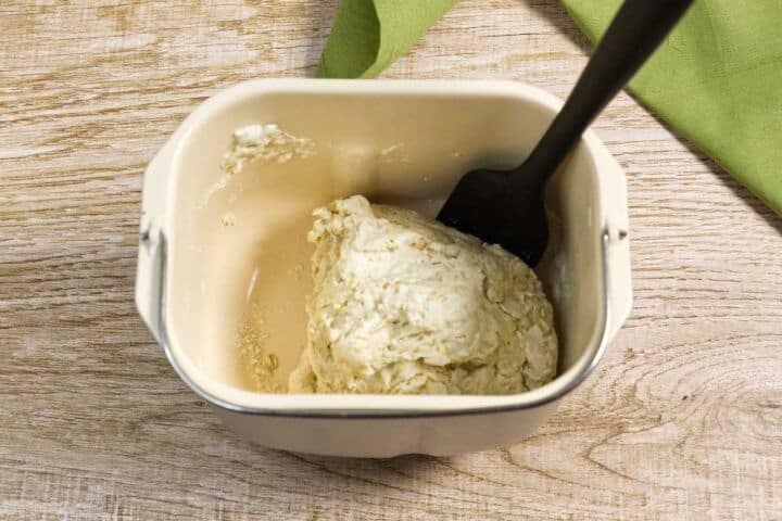 A plastic spatula rests in a bread machine pan holding a ball of raw dough, set on a wooden surface with a green cloth partially visible in the background.
