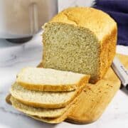 A loaf of bread machine oatmeal bread sits on a wooden cutting board, partly sliced with three pieces laid in front. A bread knife is beside the board, and a bread machine is visible in the background.