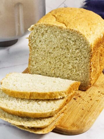 A loaf of bread machine oatmeal bread sits on a wooden cutting board, partly sliced with three pieces laid in front. A bread knife is beside the board, and a bread machine is visible in the background.