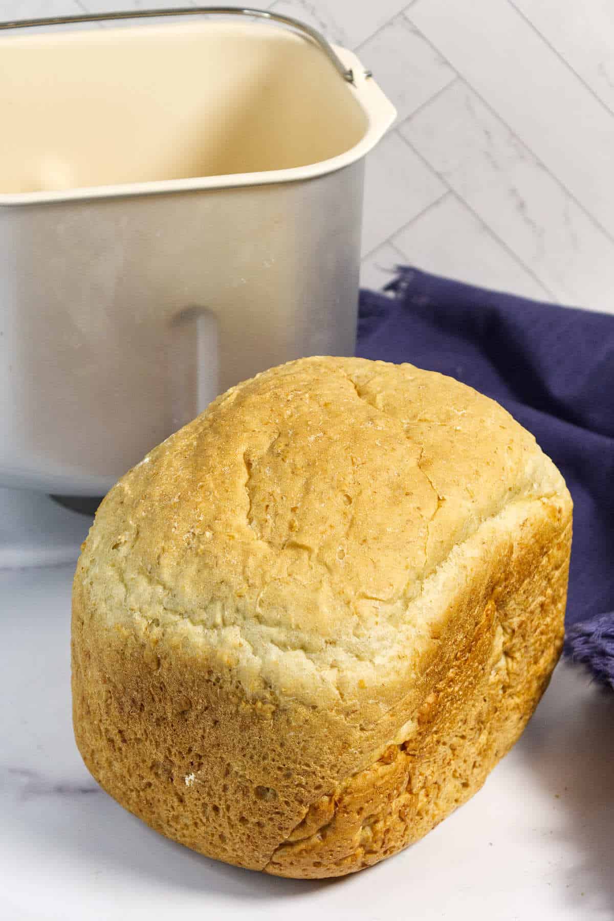 A golden brown loaf of bread sits in front of a bread machine pan, with a blue cloth nearby on a white and marble-patterned surface.