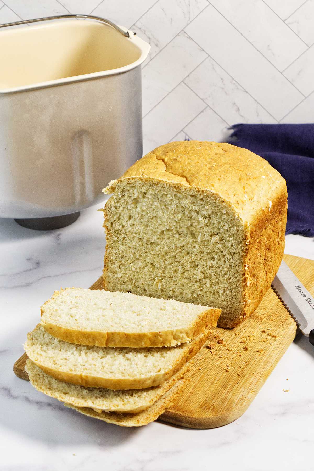 A loaf of bread machine oatmeal bread , partially sliced, sits on a wooden cutting board with three slices in front. A bread knife and a bread machine pan are nearby on a white countertop with a tiled backsplash.