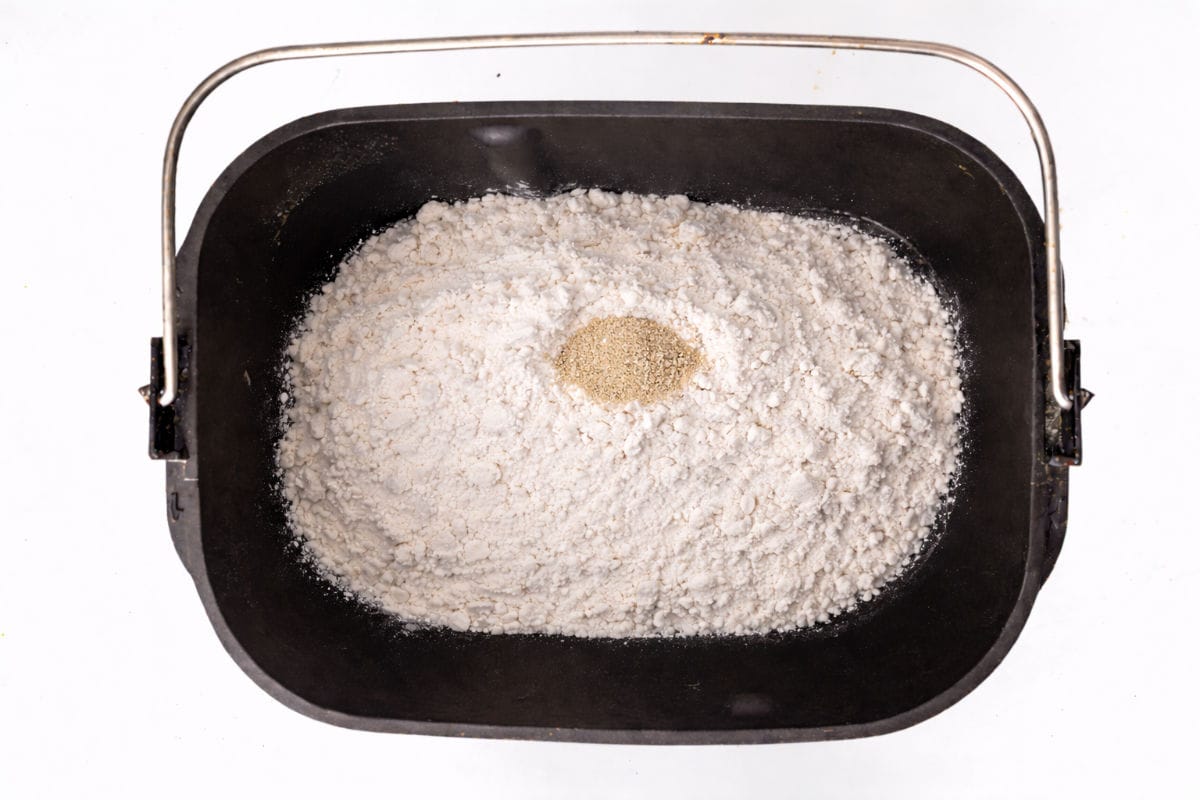 A black bread machine pan filled with flour and a small mound of yeast in the center, viewed from above on a white background.