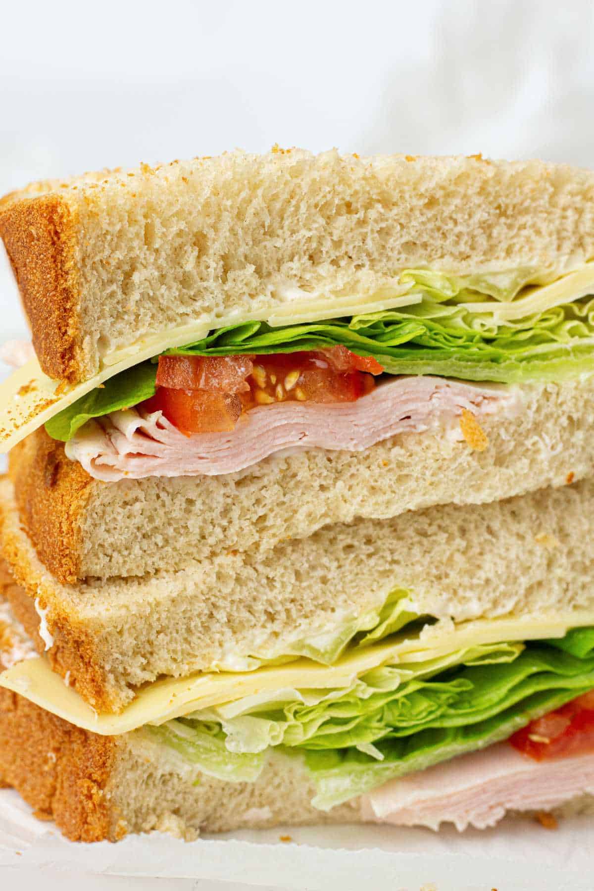 A close-up of a stacked sandwich with white bread, leafy lettuce, tomato slices, turkey deli meat, and cheese. The sandwich is cut in half and the layers are clearly visible.