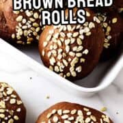 A dish of dark brown bread rolls topped with oats, with a few rolls placed on a white marble surface. The text on the image reads "Cheesecake Factory Brown Bread Rolls.