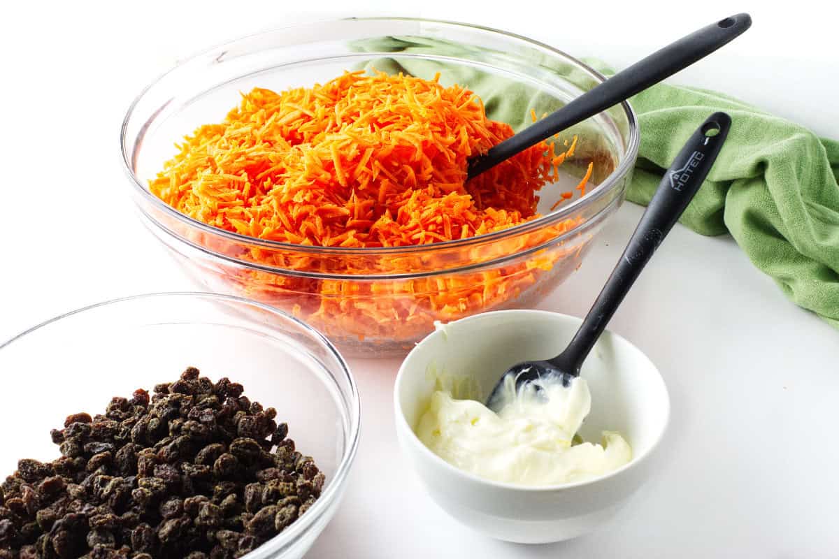 Mayonnaise dressing ingredients beside shredded carrots and raisins for carrot salad.
