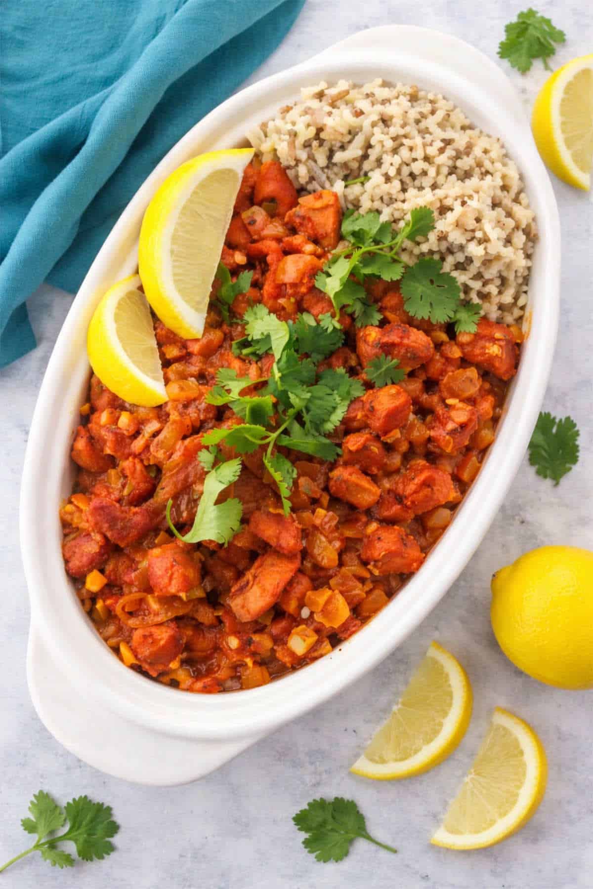 Chicken Mozambique in a white serving dish with cilantro, rice, and lemon wedges.