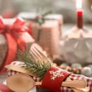 A festive table setting with gold cutlery on a red plate, wrapped in a red and white napkin and decorated with a red napkin ring and pine sprig. In the background are wrapped gifts and a lit red candle.