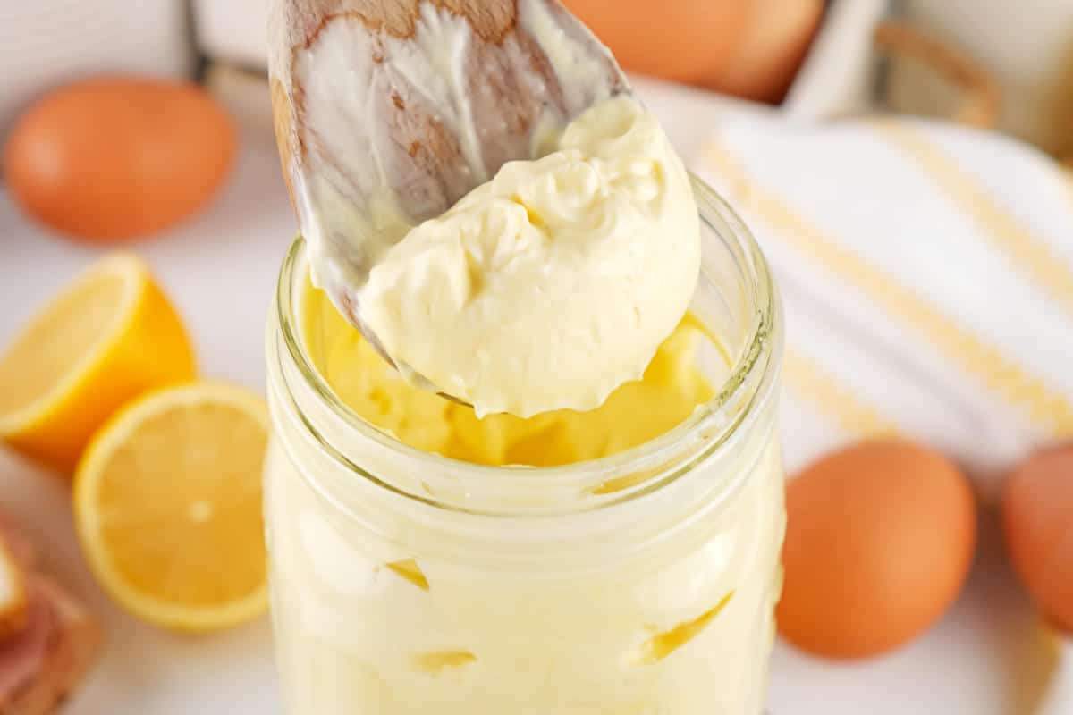 A wooden spoon scoops creamy, yellow mayonnaise from a glass jar, with fresh eggs and halved lemons visible in the background.