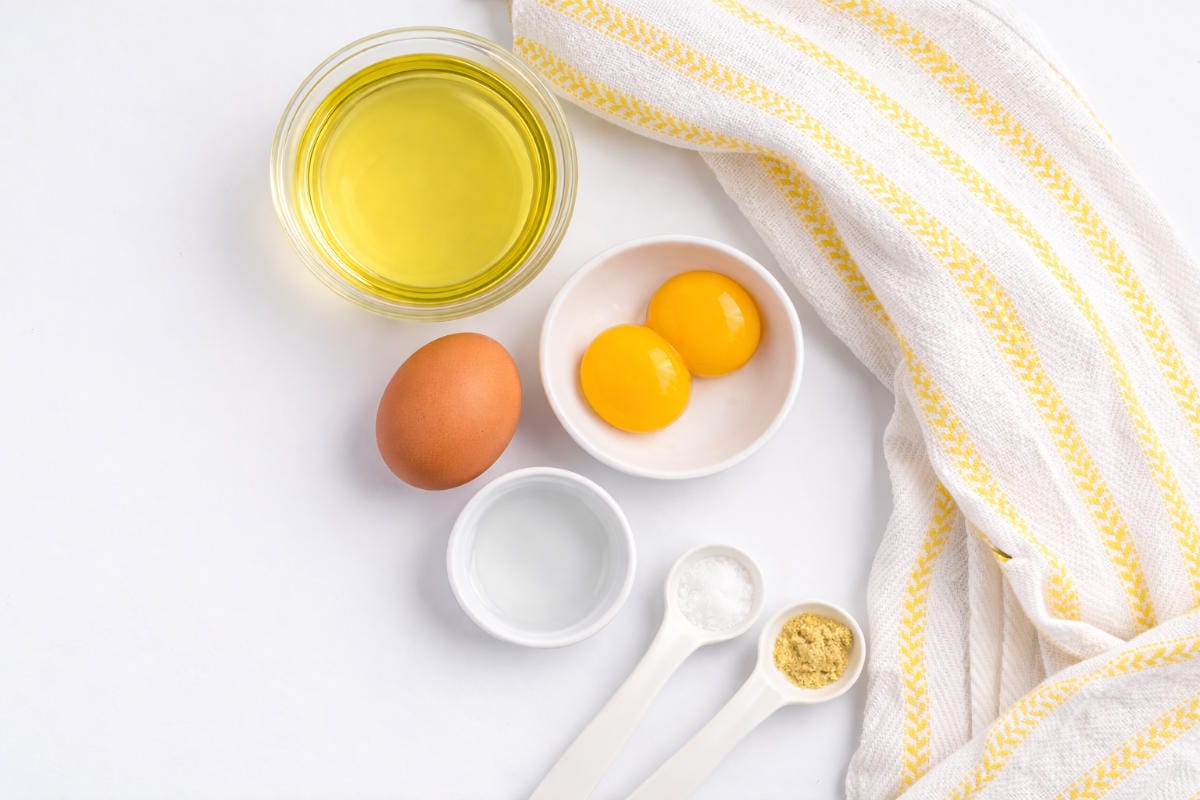 A bowl of oil, a whole egg, two egg yolks in a dish, a small bowl of liquid, three measuring spoons with powder, and a white towel with yellow stripes on a white surface.
