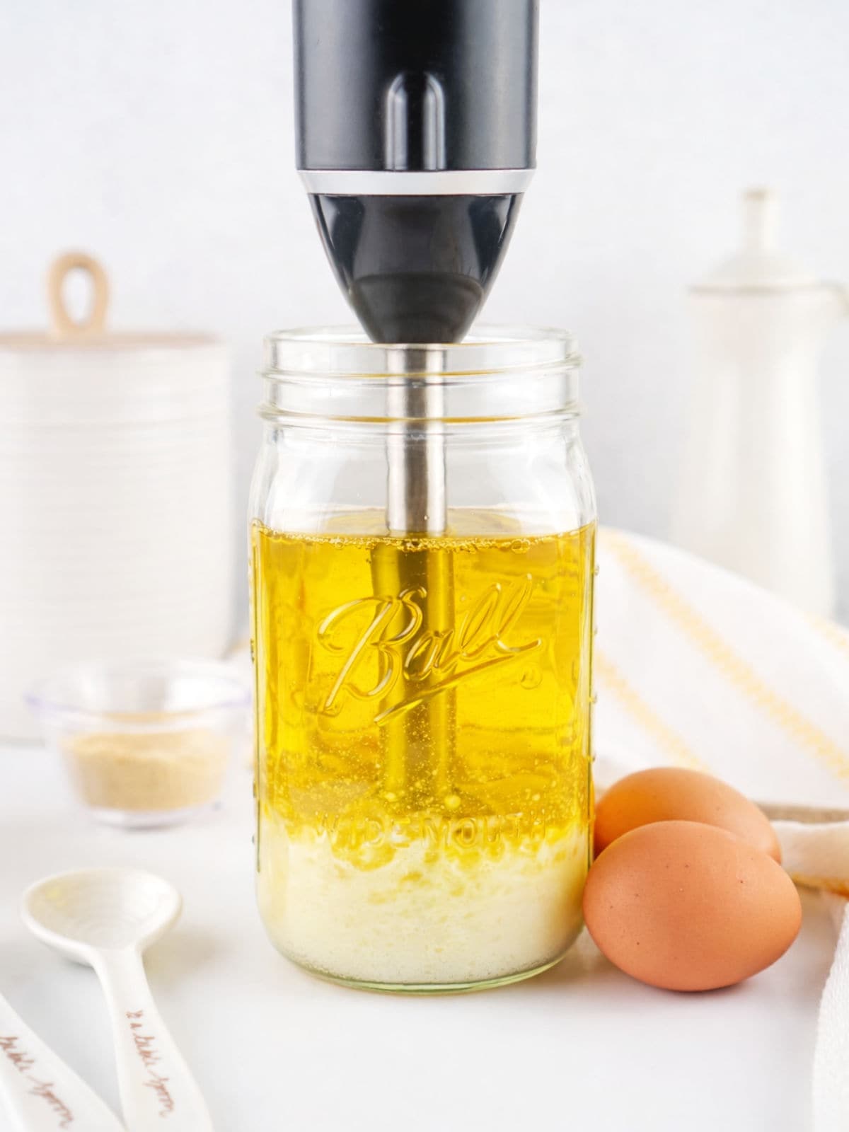 A black immersion blender is blending ingredients in a Ball mason jar filled with oil and other components. Two brown eggs, a white kitchen towel, a spoon, and a container are nearby on a white countertop.