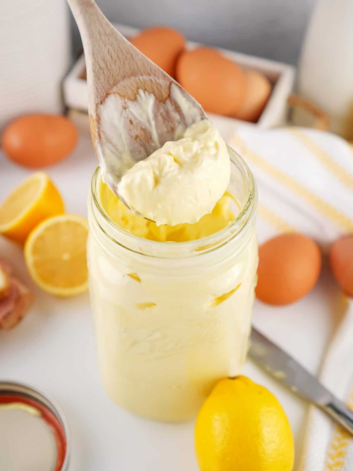 A wooden spoon scoops thick, creamy mayonnaise from a glass jar. Fresh eggs and lemons are scattered around on a kitchen counter in the background.