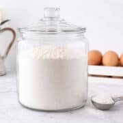 A large glass jar filled with flour sits on a marble countertop, with a metal scoop of flour beside it and a carton of brown eggs in the background.